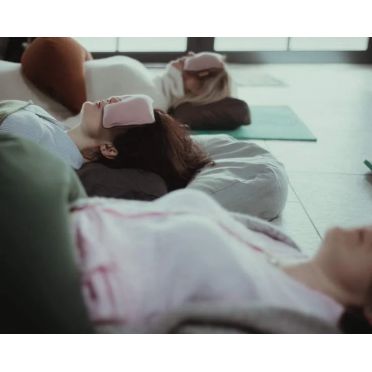 Trzy osoby leżą na plecach na macie do jogi z poduszkami na oczy w kolorze polarny róż, w tle sala z dużymi oknami.